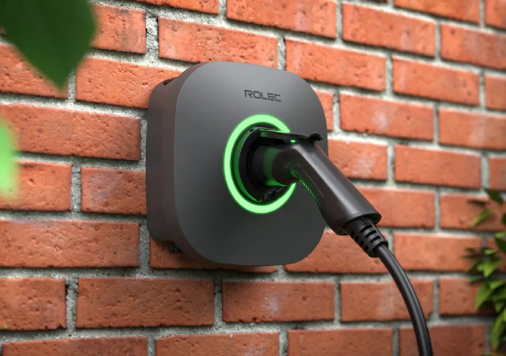 Close-up of the Rolec EVO EV charger mounted on a red brick wall, glowing green while actively charging an electric vehicle. Green leaves frame the foreground, adding a residential garden touch.